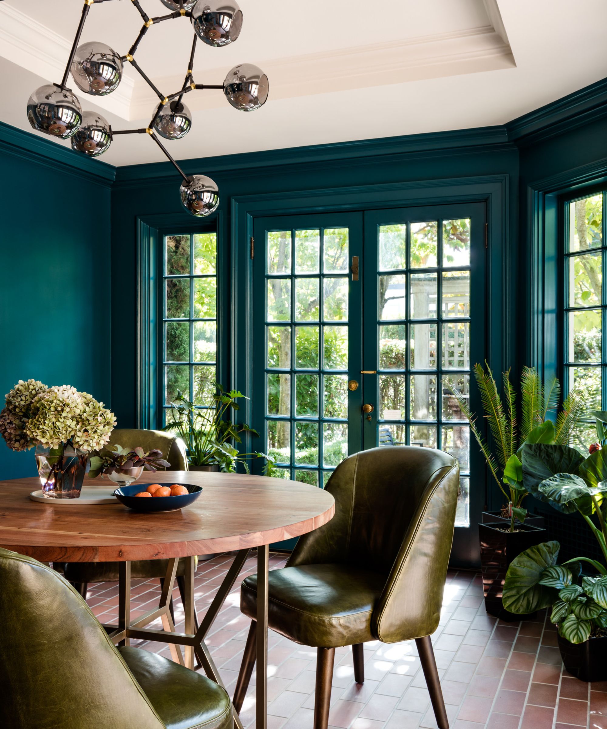 A breakfast room with terracotta-tiled flooring, teal walls and French doors leading to a garden, a white ceiling, a round wood table with green leather chairs, and a modern, sculptural ceiling light.