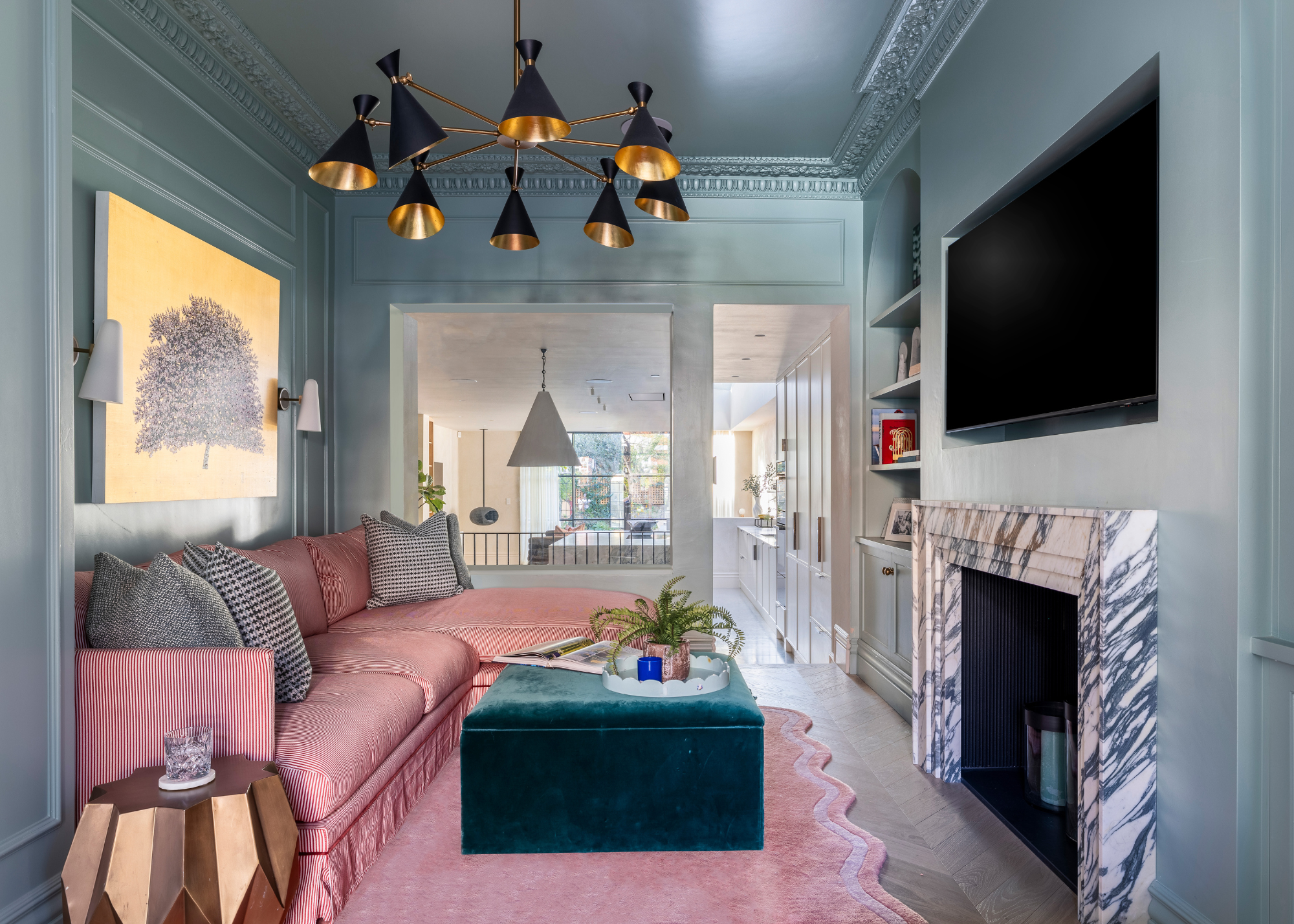 A modern living space with pink sofa and pink scalloped rug with a teal ottoman coffee table, which faces the tv and fireplace, styled with a round scalloped light blue tray containing a fern and a blue candle next to an open book