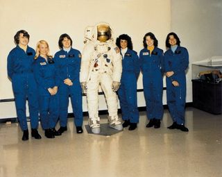 six women in blue flight suits, standing three on each side of a spacesuit mannequin