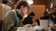 Student working on a laptop computer in a university library, with other students working at desks in background.