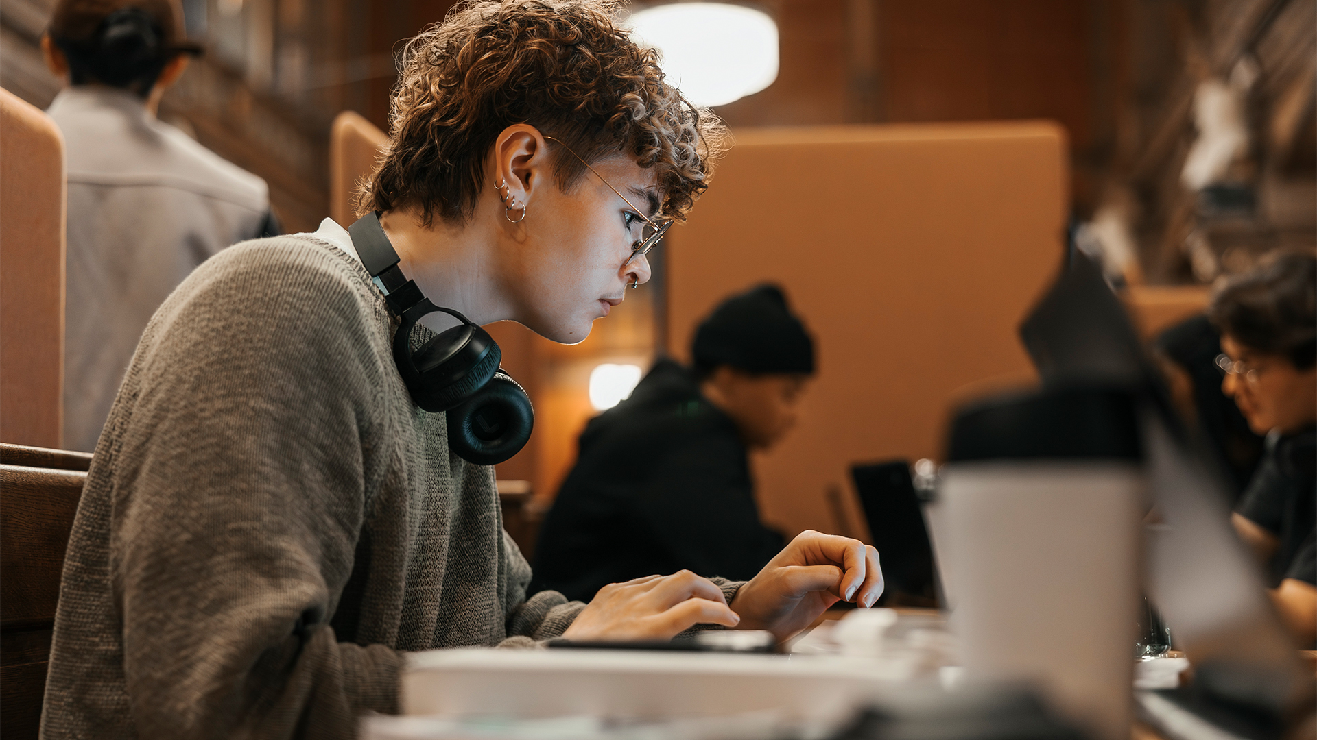Student working on a laptop computer in a university library, with other students working at desks in background.