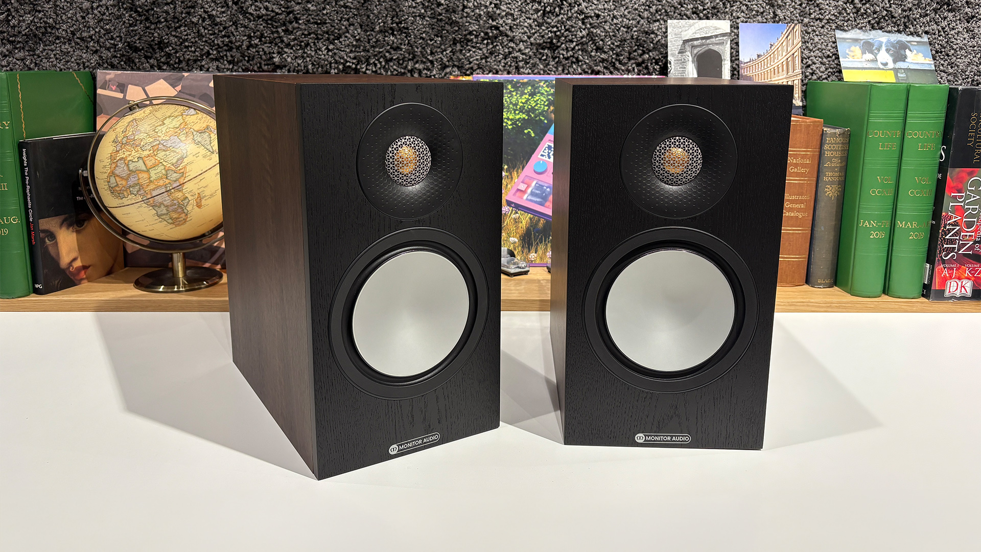 Monitor Audio Bronze 50 7G standmount speakers on white surface in front of busy bookcase