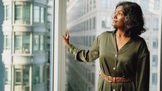 An older businesswoman wears a thoughtful expression as she stands by a window in a high-rise and looks outside.