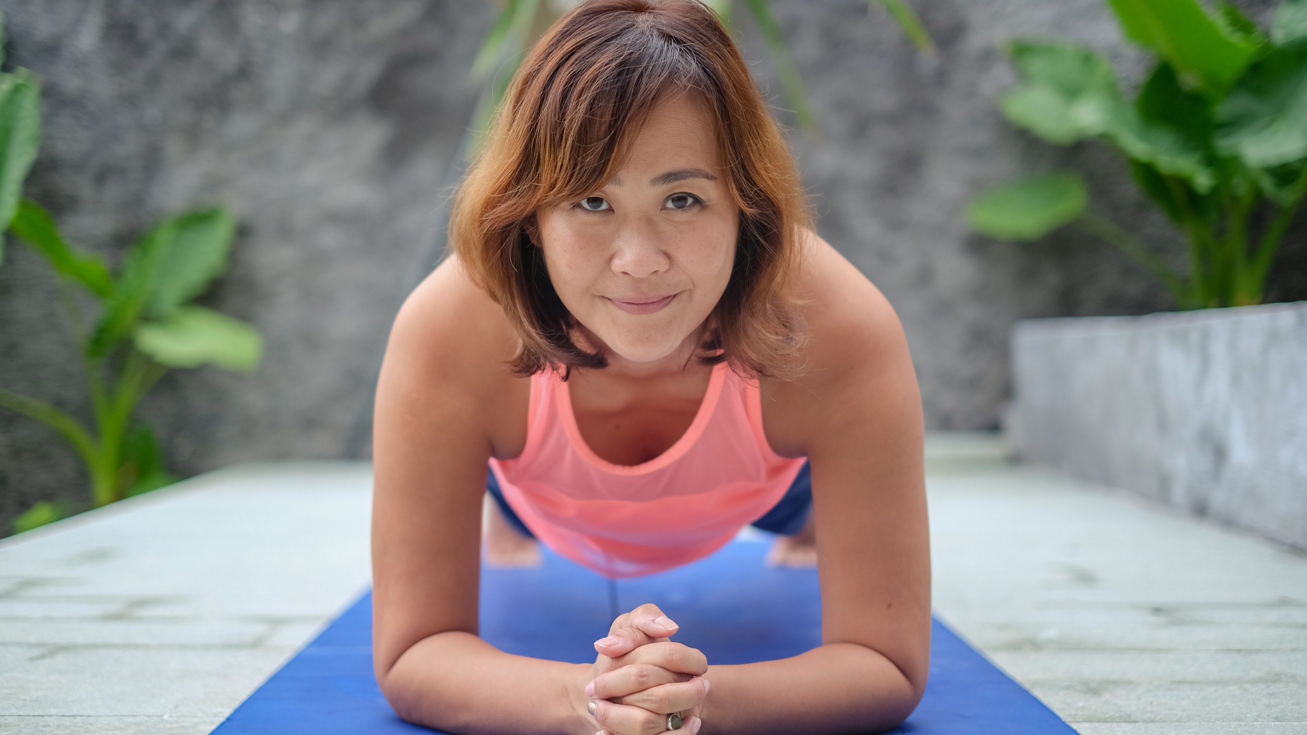 A woman smiles at the camera as she performs a plank exercise on a blue yoga mat.