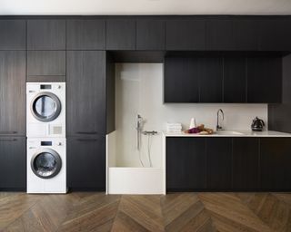 Wall to ceiling laundry room storage ideas in a space with black wood ceiling height cabinets, a dog washing station and stacked washing machine and tumble dryer on wooden flooring.