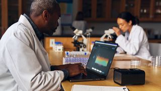 A man wearing a white lab coat sits in front of his laptop inside of a science laboratory. A woman in the background, wearing a white lab coat, looks into a microscope.