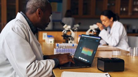 A man wearing a white lab coat sits in front of his laptop inside of a science laboratory. A woman in the background, wearing a white lab coat, looks into a microscope.