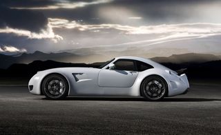 Side view of Weissmann’s purist sports car in white, grey road, grass verge and silhouette mountainous landscape, dark grey cloudy sky