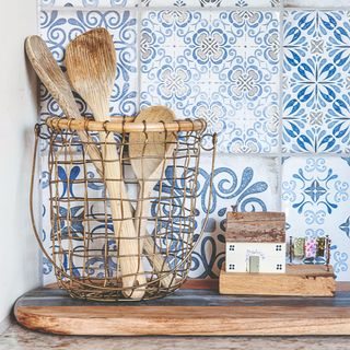 Wooden chopping board and basket of wooden spoons in front of a grey, white and blue tiled wall