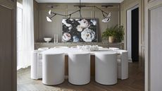 Dining room with white upholstered chairs and wood and white table, branched light above, and sideboard with large floral artwork, plant, bowl and candlesticks