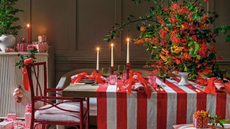 A Christmas table decorated with a red and white striped tablecloth, lit taper candles in varied height red taper candleholders, red napkins and a vase with oversized colorful foliage