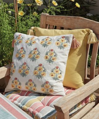 Two throw pillows - one yellow and one white with yellow bouquet patterns - on a wooden garden chair with a pastel colored seat cushion with a striped dining table in front of it and grass with yellow wildflowers