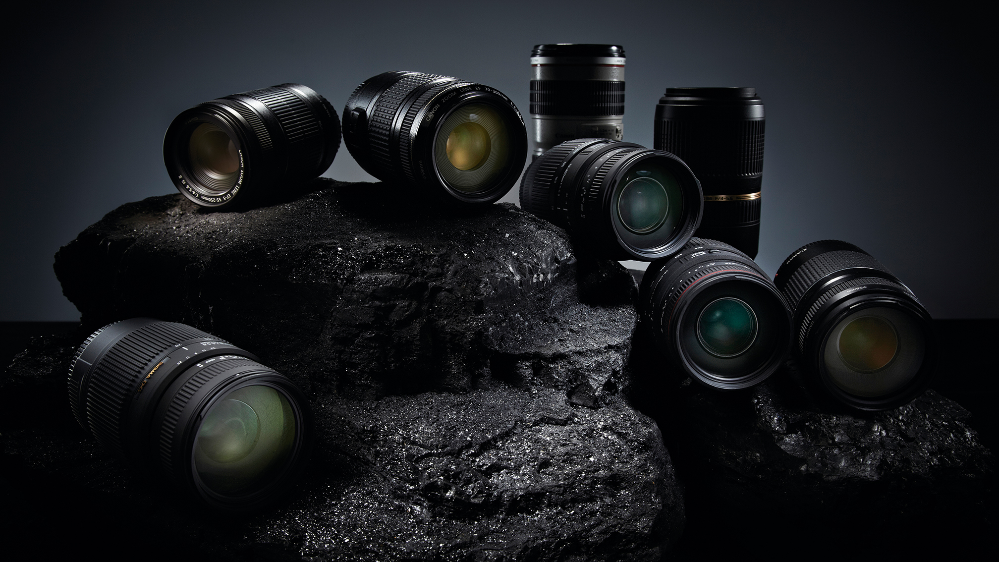 Eight assorted camera lenses arranged on a dark, textured rock surface with dramatic lighting