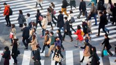 Image of a crowd of people crossing the Shibuya intersection. One set of white marking lines can be seen running parallel to the bottom side of the image, while another run diagonally towards the upper left-hand corner of the image. A range of people can be seen crossing from those carrying backpacks, others carrying shopping bags, some dressed in suits and others in jeans