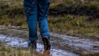 A pair of legs on a muddy trail, wearing hiking trousers and a pair of Salewa Mountain Trainer Mid Gore-Tex Boots.