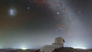 The domed roof of the Vera Rubin Observatory sits on a high ridge with a red and purple starry night sky above it with a glowing arch of the Milky Way seen in the heavens