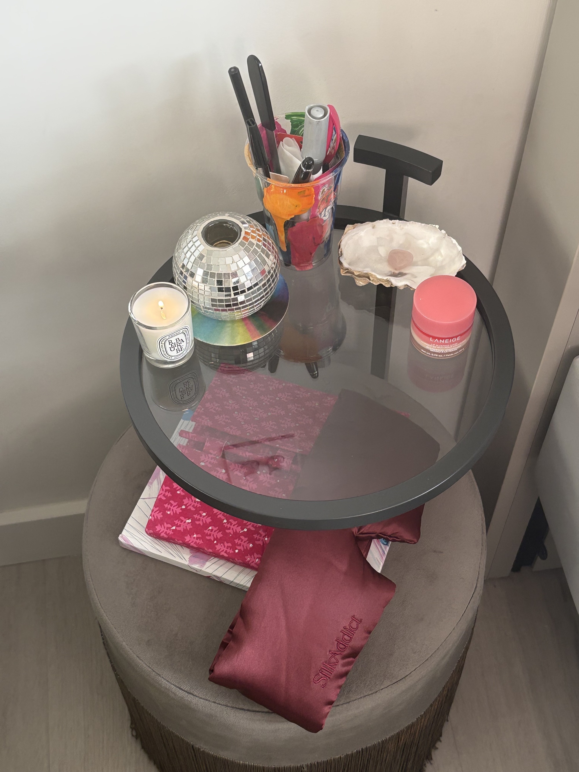 Image of a gray, two-story nightstand in the corner of a room. There is a cup of pens, a mini disco ball, a candle, and a large eye mask on it.