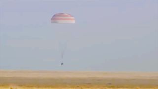 a cone-shaped capsule descends onto flat grassland under a parachute