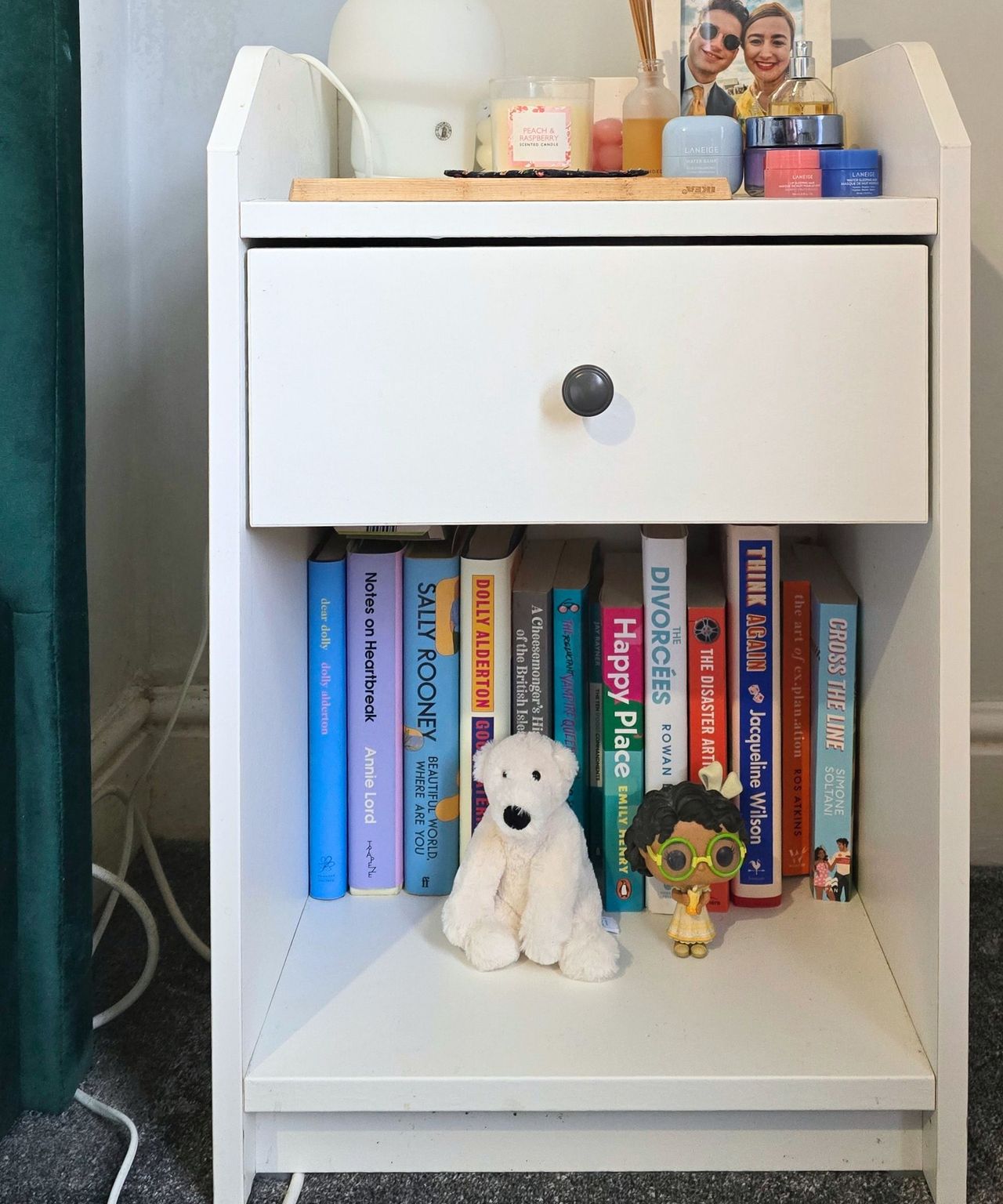 A white nightstand with colorful books, a polar bear toy, and a Funko Pop on the bottom shelf, and makeup tubs, a lamp, and a photo frame on the top shelf