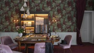 Burgundy wallpaper with green leaves on it in a dining room setting with velvet chairs and a large dining room table