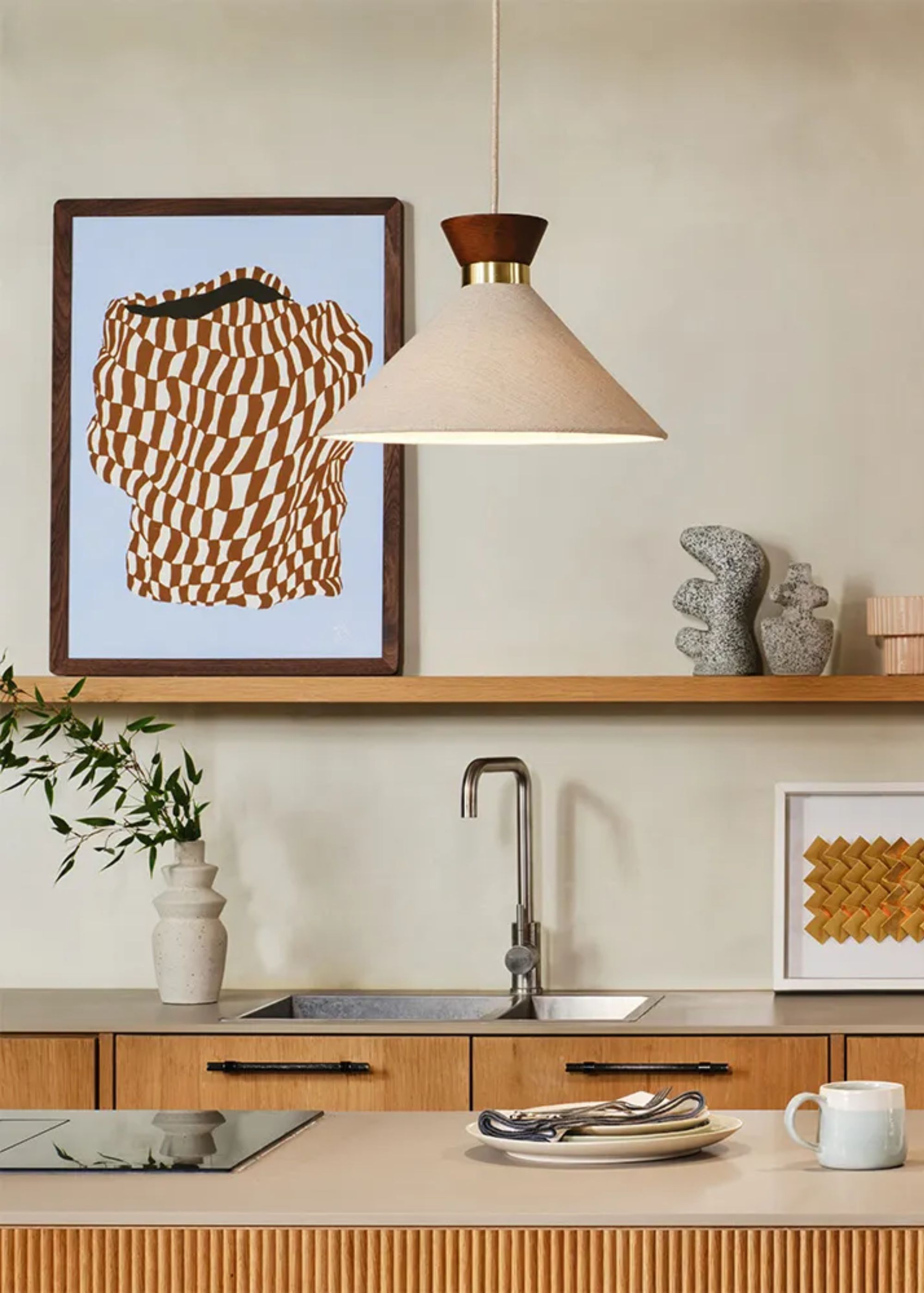 A kitchen with a large shaded pendant light hanging above it. There is also an open shelving area in the kitchen with artwork placed on top of it. On the island is a plate and a mug