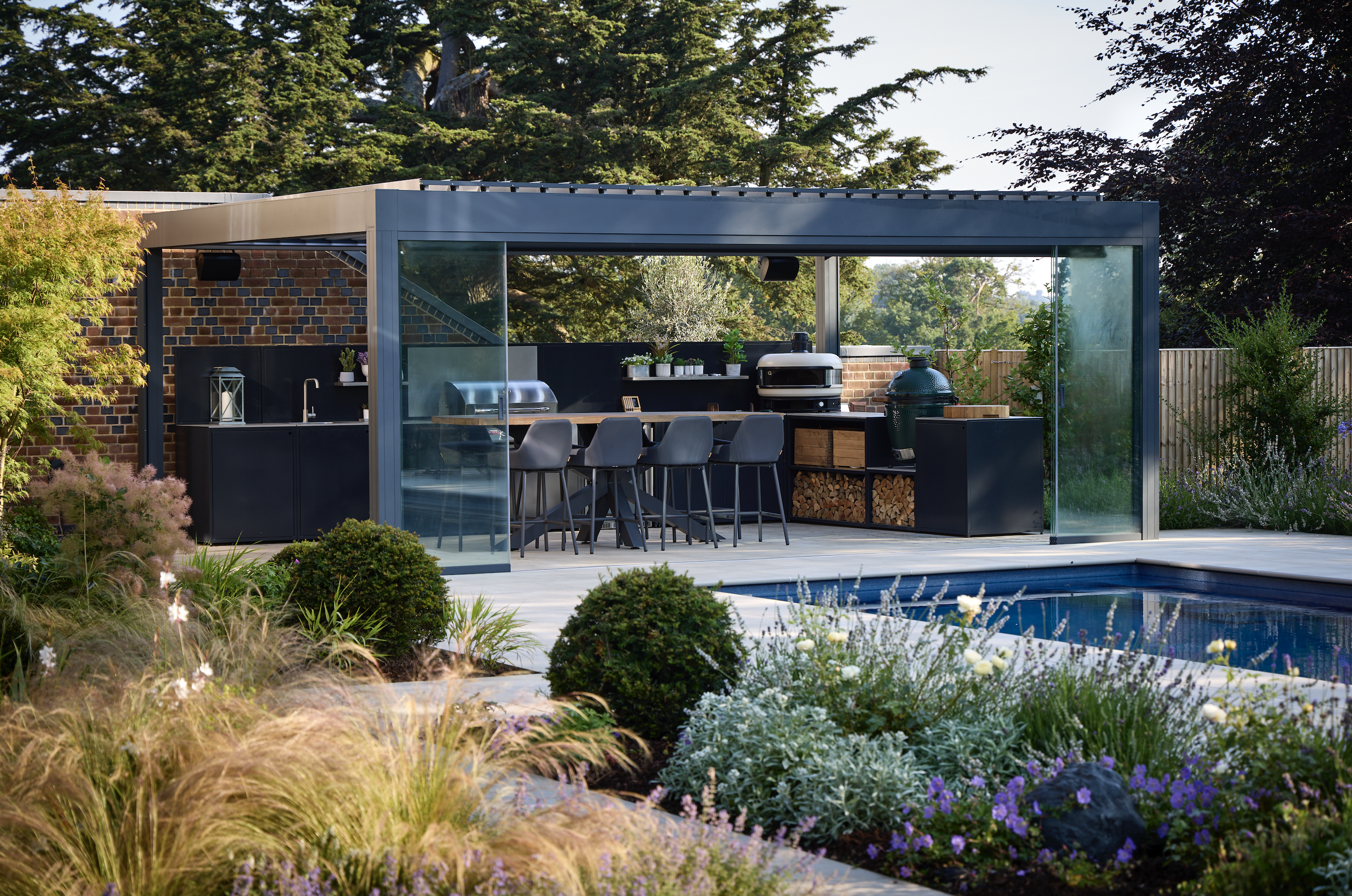 An outdoor kitchen set inside a pergola, with dining chairs and built in grills