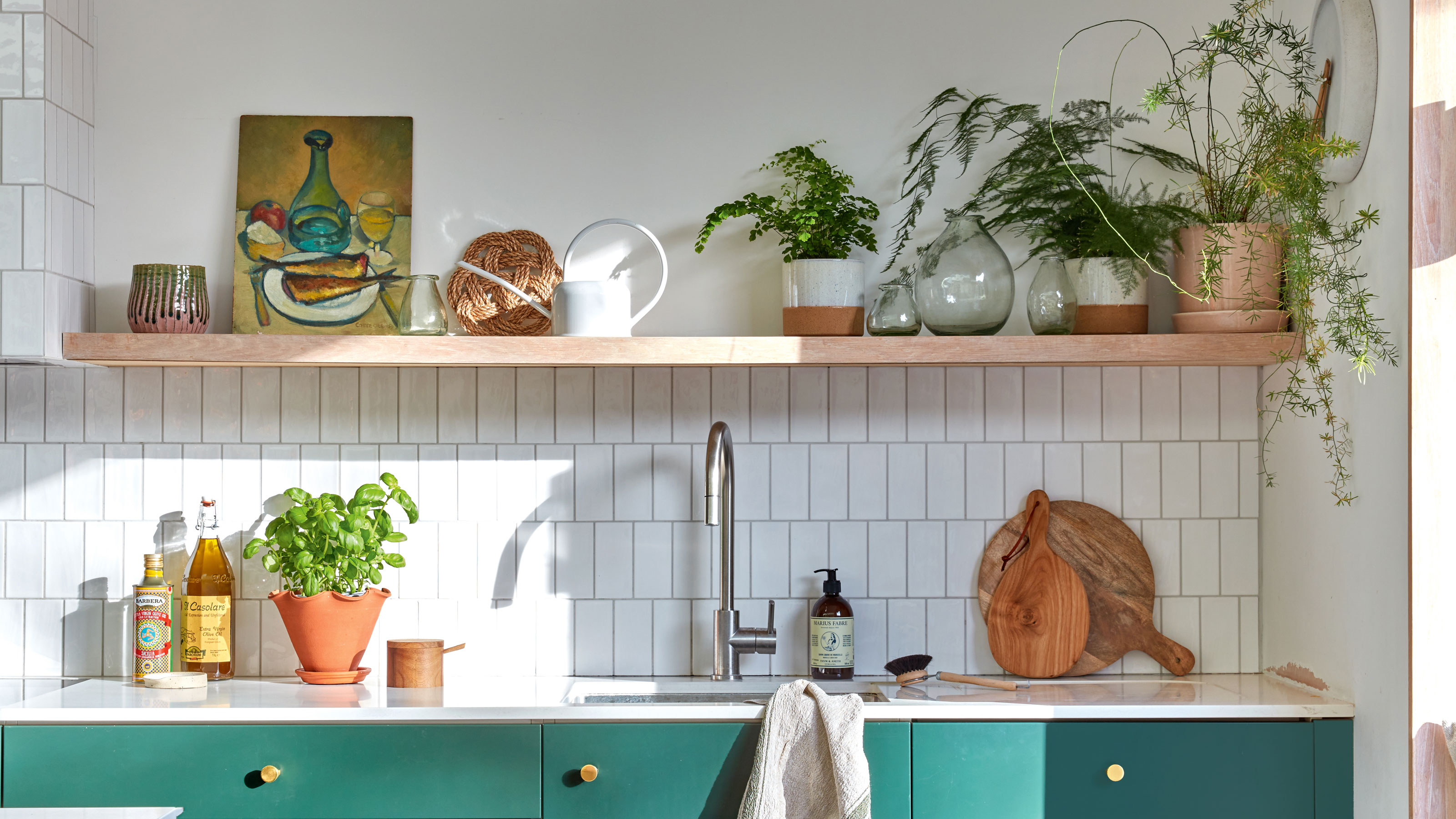 White kitchen tiles with green cabinets and wooden open shelving over a sink, with a pot of basil on the counter.