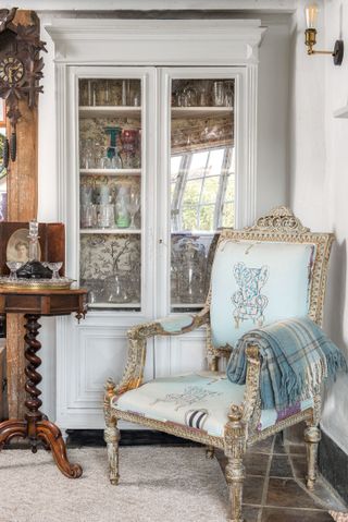 painted glazed cabinet with glassware and gilded armchair with blue upholstery and dark wood table with turned pedestal