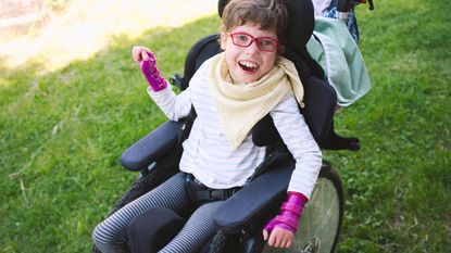 A smiling girl with purple arm braces sits in a wheelchair.