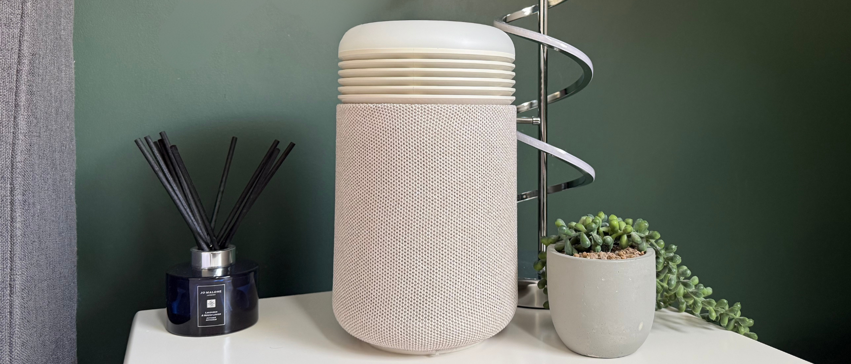 Blueair Mini Restful sunrise alarm clock and air purifier on a bedside table next to a lamp, plant and reed diffuser, showing its compact design