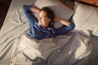 Image depicts someone laying in bed wearing striped pyjamas. They have a smile on their face and are wearing the Nurosym device in their left ear