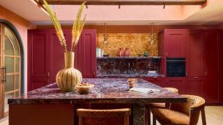Image of a large red kitchen with a large red marble island that has orange tiger-print bar chairs around it. There is a large yellow vase with feathers, pink ceilings, and a skylight.
