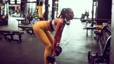 woman in a gym wearing yellow leggings and white crop top holding two dumbbells performing an RDL exercise. 