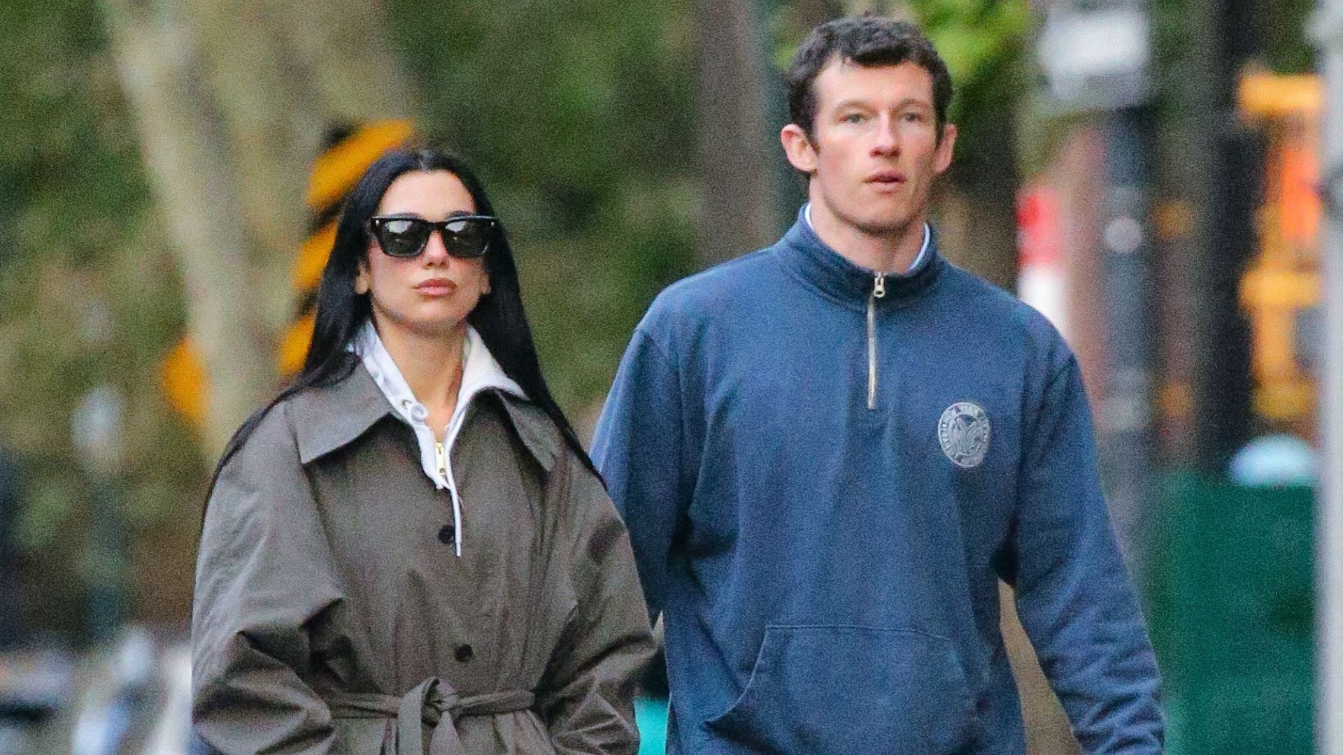 Dua Lipa walks down the street with Calum Turner wearing a grey single-breasted trench coat with leggings, a hoodie and black trainers.