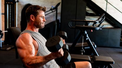 Chris Hemsworth performing incline biceps curls with dumbbells, seen from the side. A treadmill is in the backround