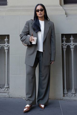 A woman standing on the sidewalk holding coffee while wearing brown sunglasses, a boxy white t-shirt, a gray boxy blazer, gray trousers, brown and white pointed heels, and a brown Gucci bag.