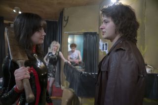 Chrissie Hynde (Sydney Chandler), wearing a black and red rubber outfit, holds a cricket bat over one shoulder while confronting Steve (Toby Wallace) in the SEX punk fashion shop run by Vivienne Westwood (Talulah Riley) and Malcolm McLaren (Thomas Brodie-Sangster), who can be seen out of focus in the background, standing by the changing rooms and watching from a distance