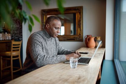 Mature cafe owner sitting at table by bright window in cafe using laptop looking at plans.