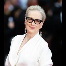 Meryl Streep wearing glasses and clear dangly earrings and smiling on a red carpet.