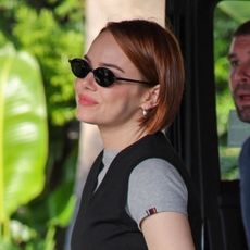Emma Stone wears black parachute pants, black strappy heels, a black preppy vest, a gray t-shirt, and carries a Louis Vuitton bag