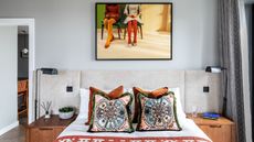 A maximalist bedroom. A double bed with colorful throw cushions on it. Two wooden nightstands with matching black lams either side. A framed photo of two people sitting down on the wall behind.