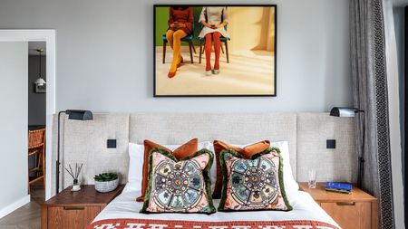 A maximalist bedroom. A double bed with colorful throw cushions on it. Two wooden nightstands with matching black lams either side. A framed photo of two people sitting down on the wall behind. 