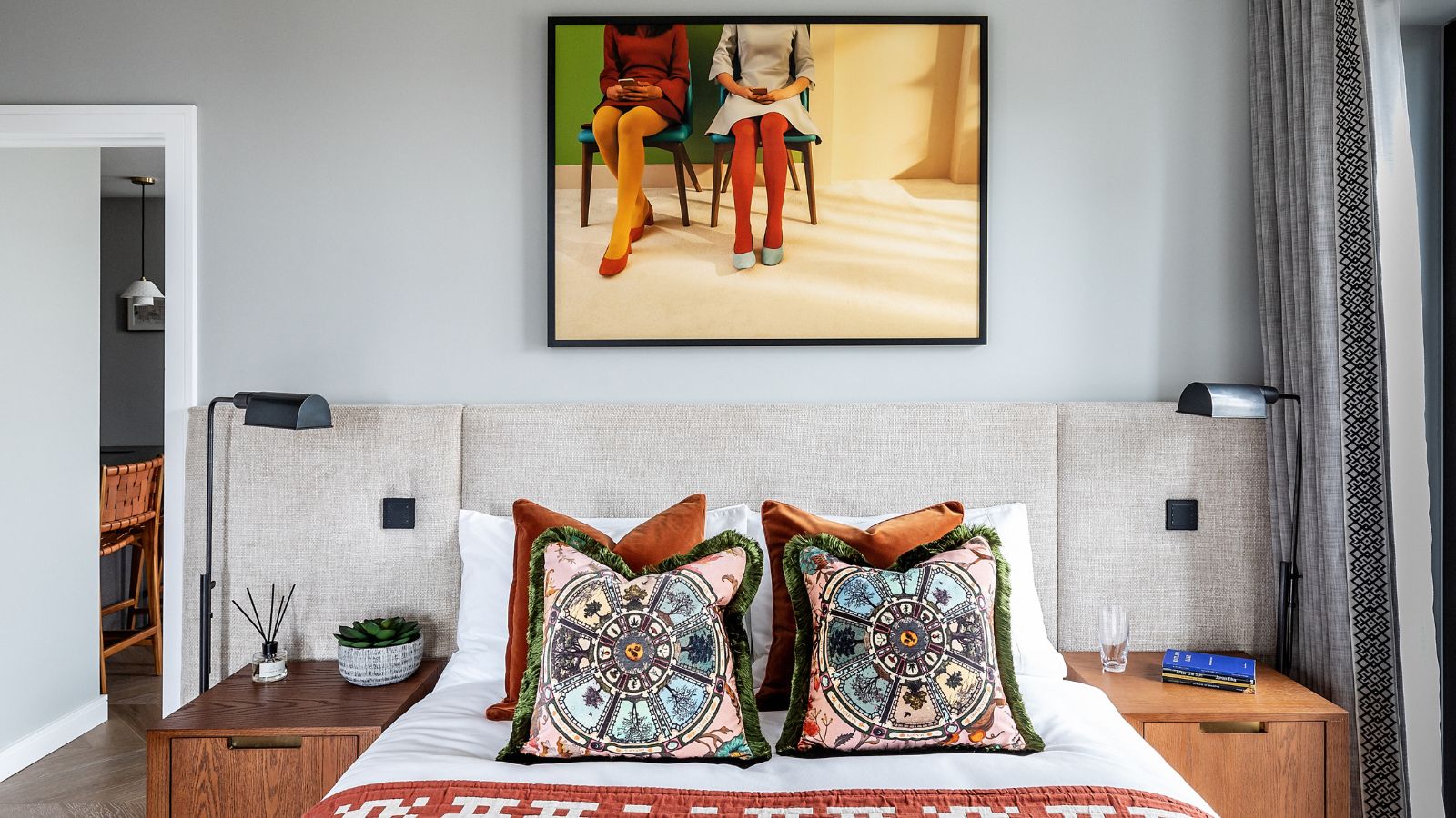 A maximalist bedroom. A double bed with colorful throw cushions on it. Two wooden nightstands with matching black lams either side. A framed photo of two people sitting down on the wall behind. 