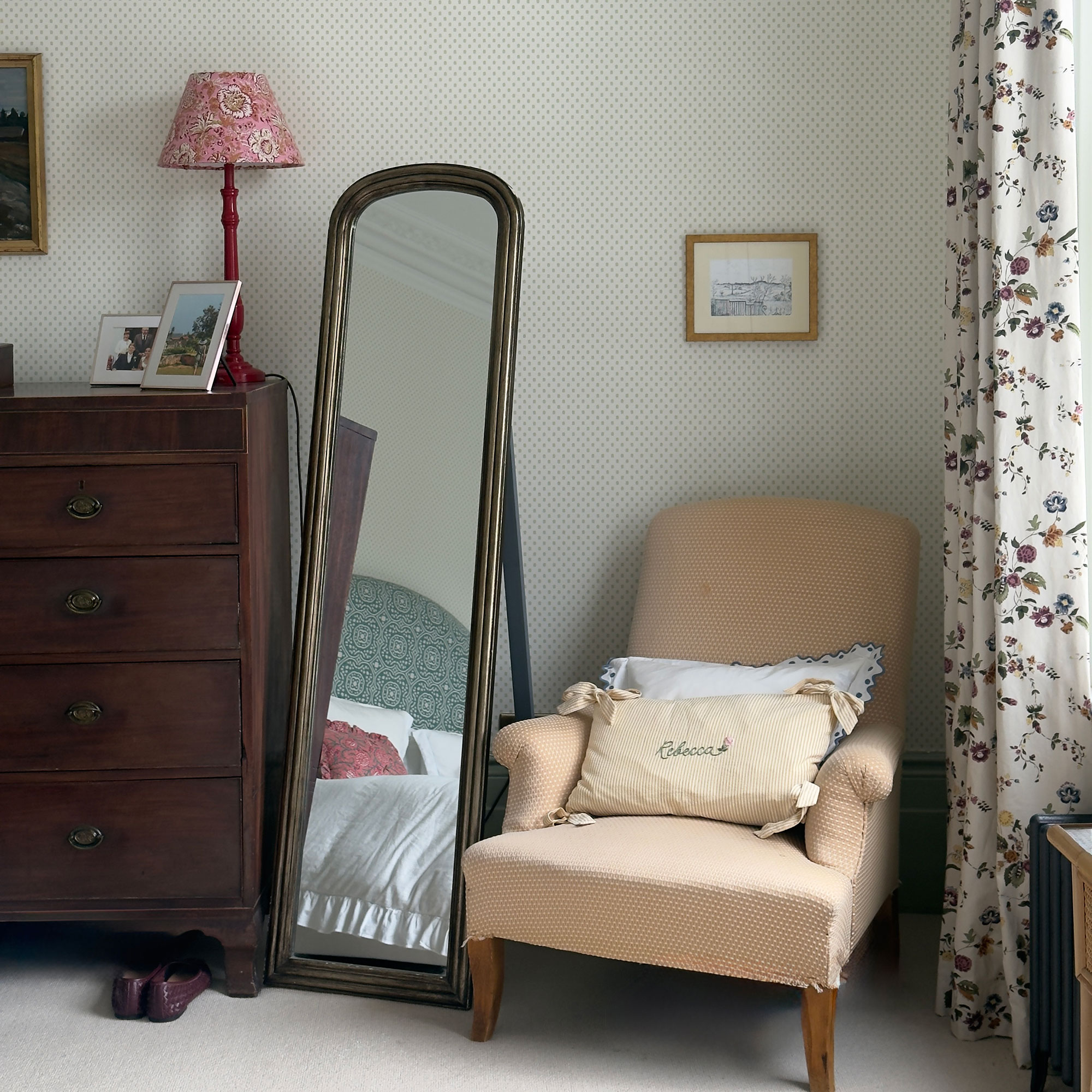 Upholstered armchair beside full length standing mirror and dark wood chest of drawers in corner of room