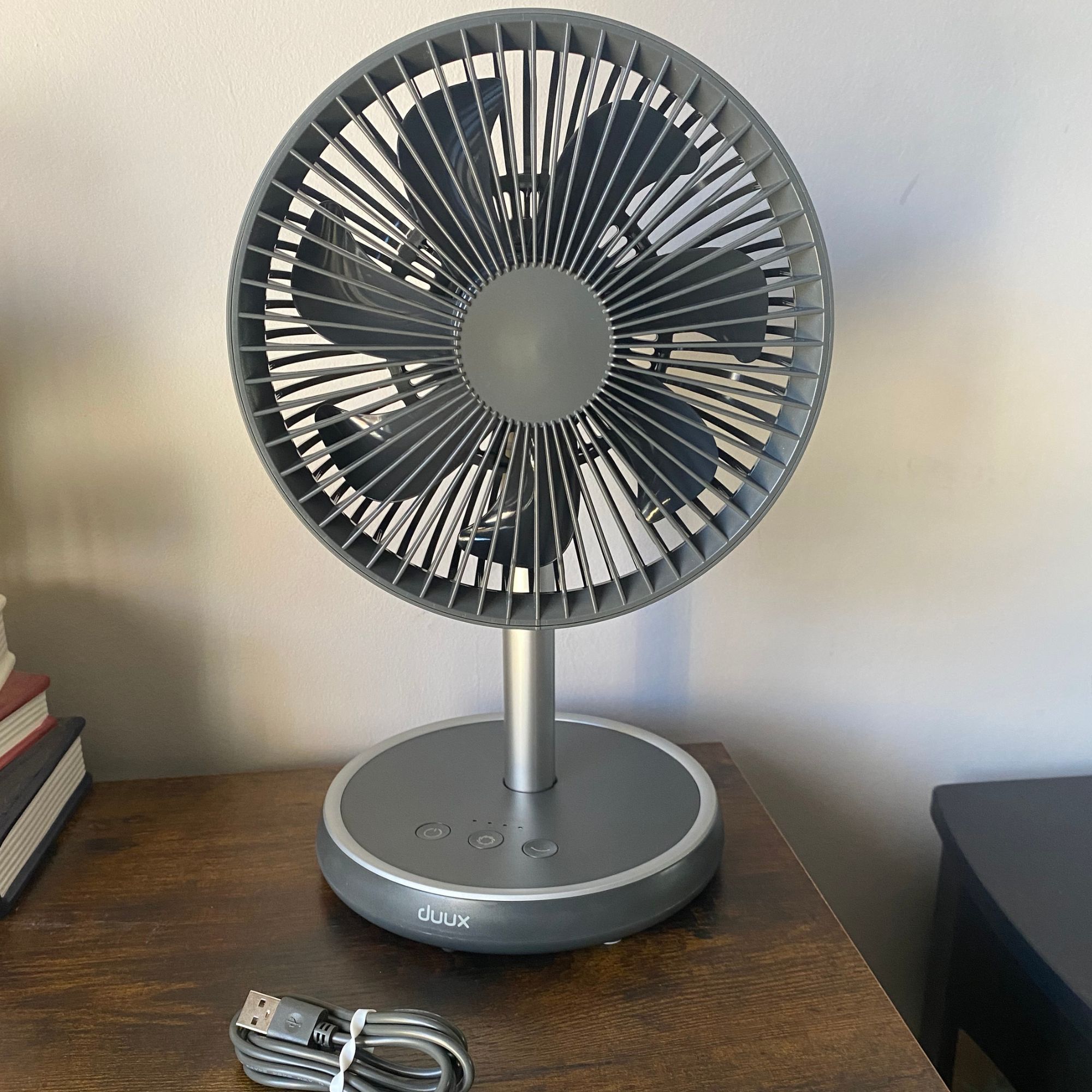 The Duux Rize fan in table top mode, with the charging cable it comes with, on a wooden top sideboard