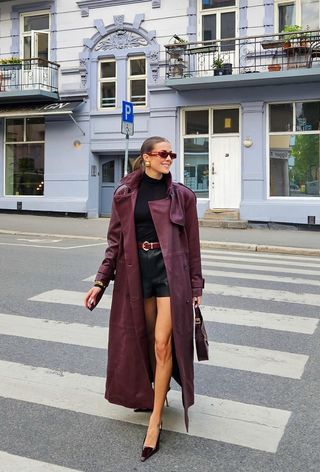 A woman wearing a burgundy red leather trench coat over a black turtleneck tucked into black leather shorts, red pumps, a red belt, and a red bag.