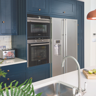 Kitchen with white walls and counters, blue cabinets, silver fridge, and sink and white worktops. Pub Orig. A four bedroom renovated Edwardian terraced house with a small extension in York, home of Philippa and James Beaumont.