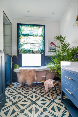 Blue and green bathroom with patterned floor tiles, a botanical blind, powder pink bathtub, fresh houseplants, and blue paneling on walls.