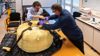 Two people wearing glasses work above what looks like a large tire with fabric on the outside on a table. 