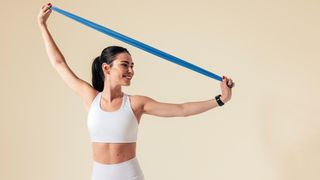 a woman wearing white workout wear stretches a resistance band above her head